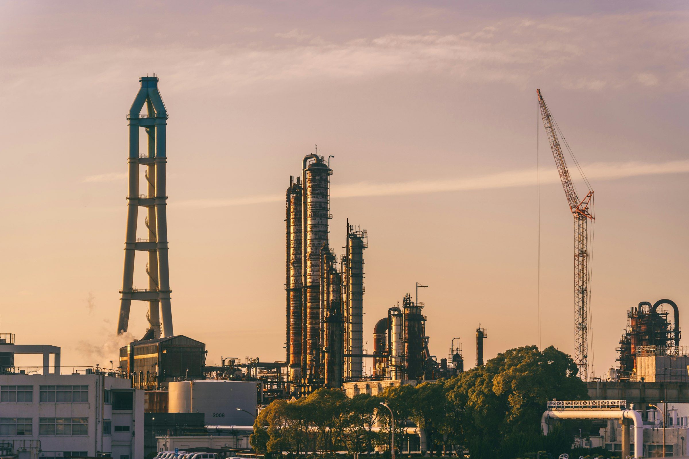 Distillation towers and stacks of an industrial processing plant at sunset, with a construction crane standing beside them against an orange sky