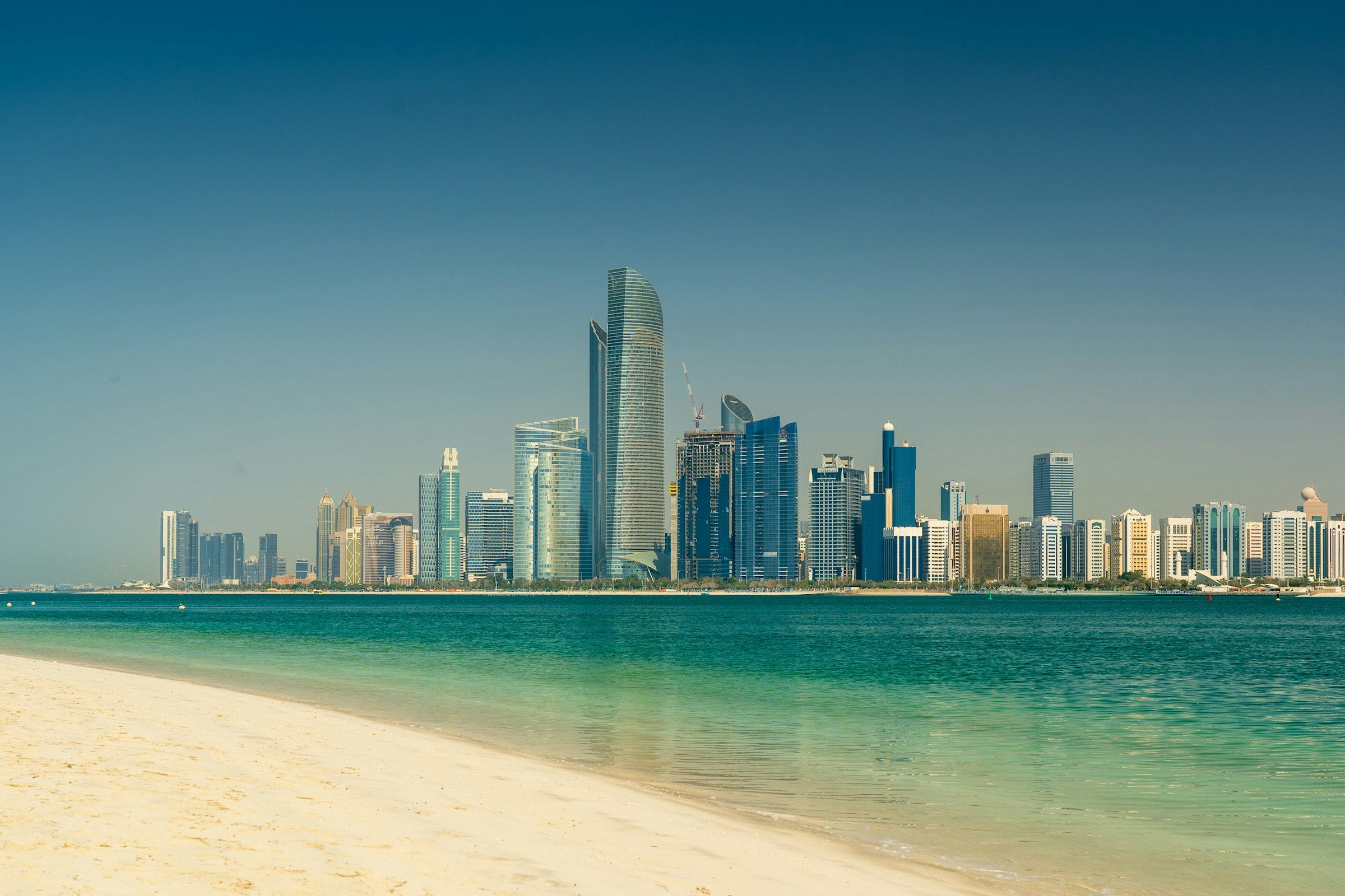 Abu Dhabi's high-rise skyline seen from a sand beach across turquoise water