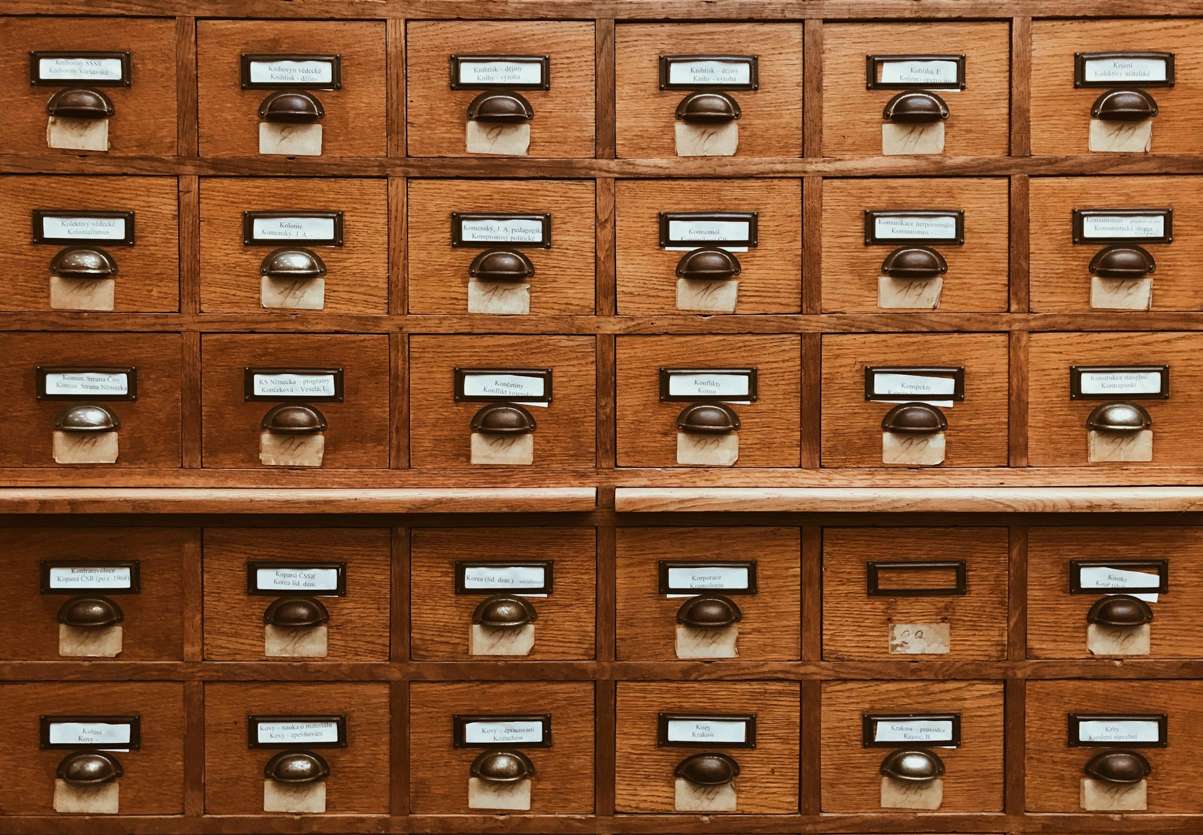 A wall of wooden library card-catalogue drawers with brass cup handles, each drawer fronted by a small typed index label