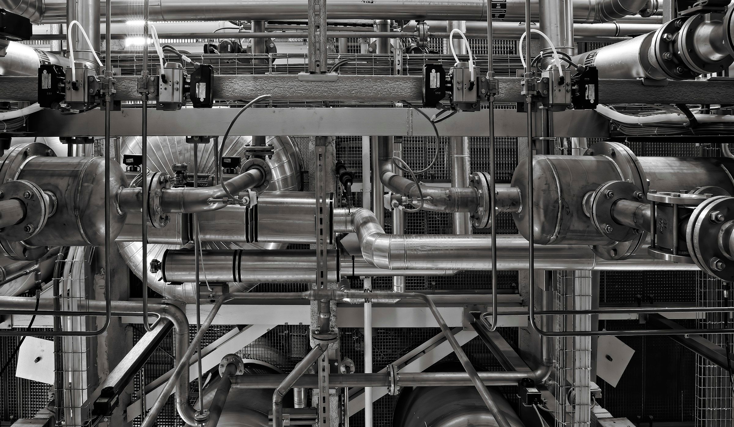Dense insulated pipework, valves and pressure vessels inside an industrial plant room, in black and white