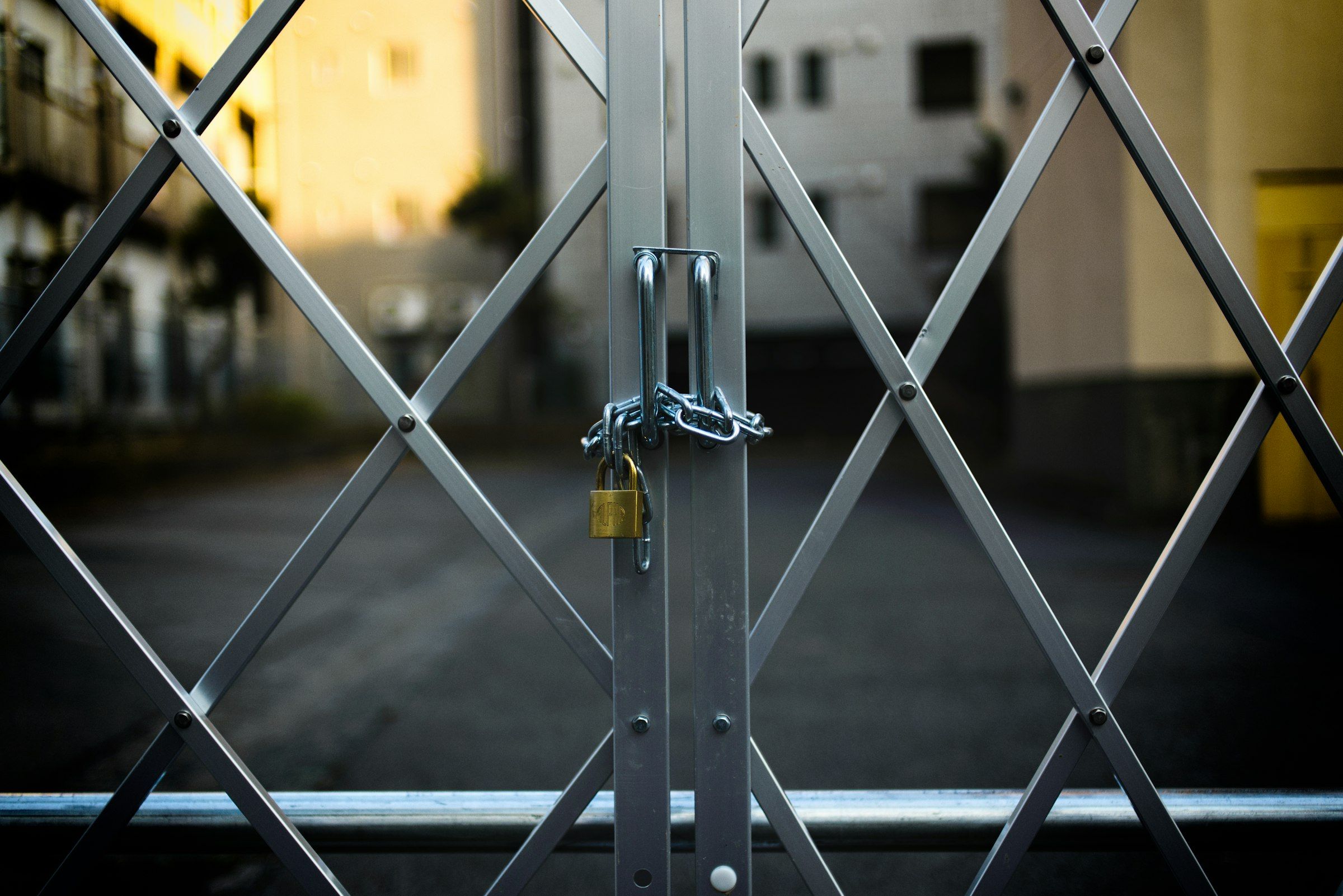 A metal lattice gate closed and fastened with a chain and brass padlock, a street out of focus behind it