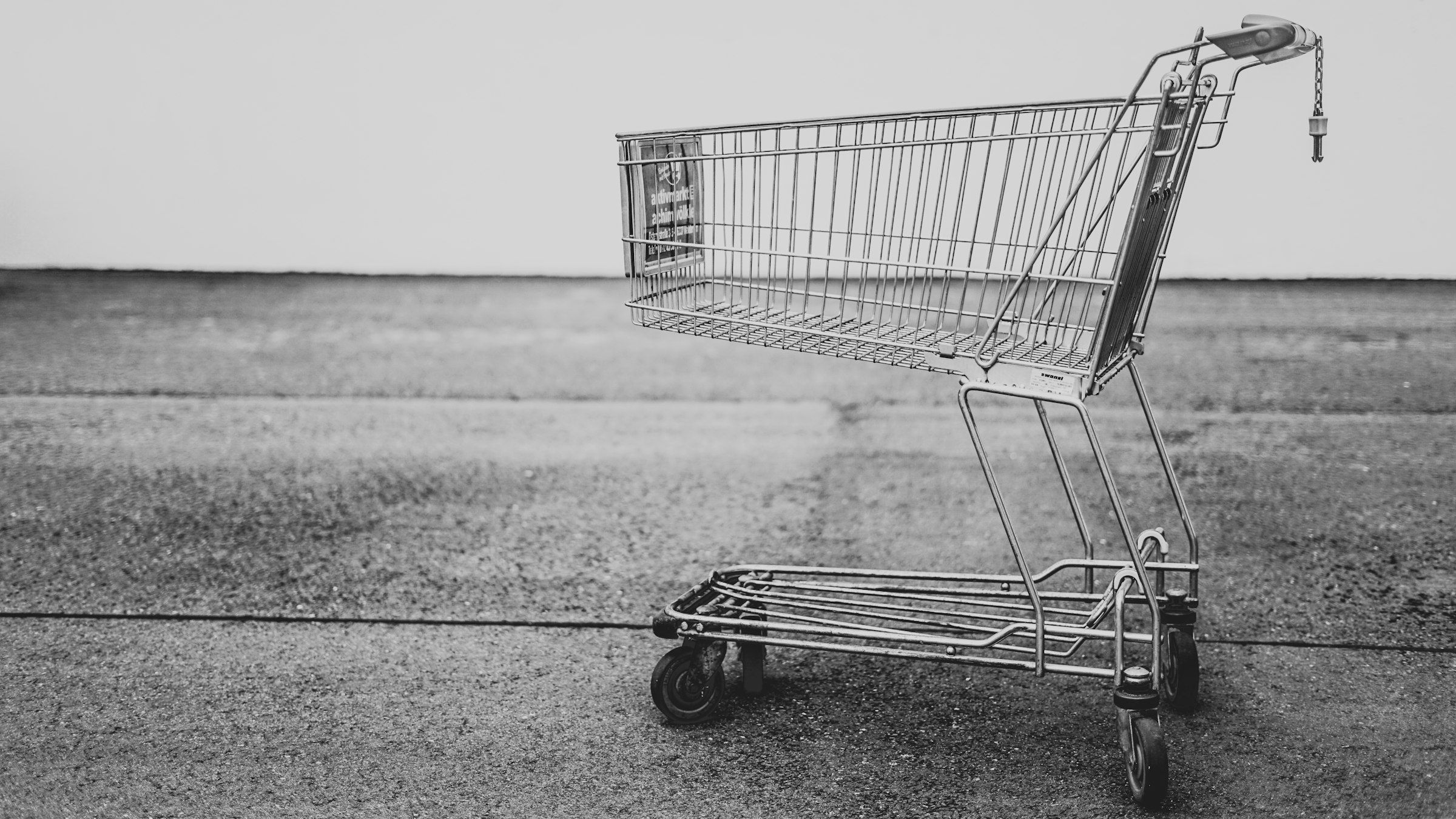 A single empty wire shopping trolley standing alone on bare asphalt against a plain pale wall, photographed in black and white
