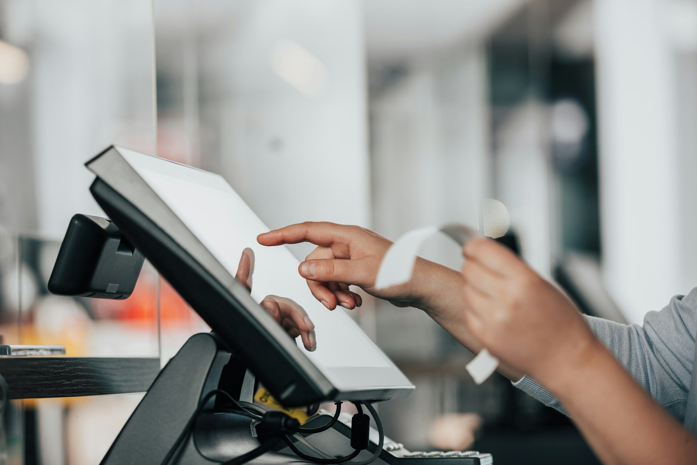 Two hands at a shop counter, one touching a blank point-of-sale screen and the other holding a curling paper receipt