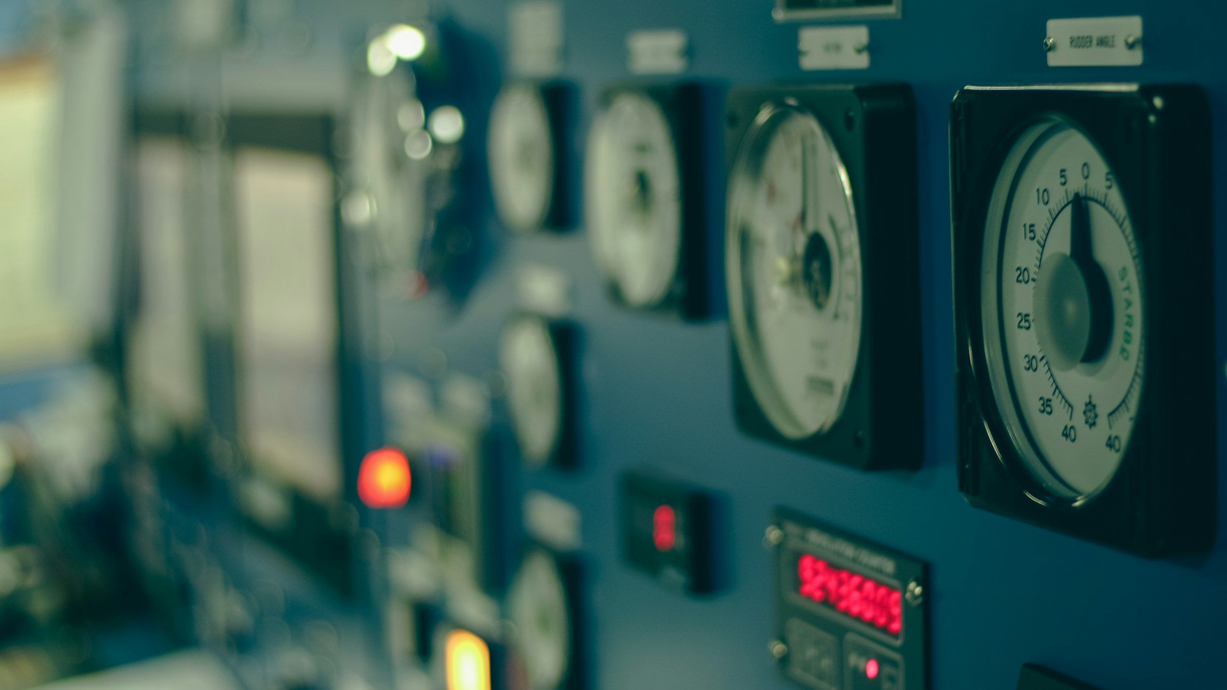 A receding wall of round analogue dials and small red indicator lights on a blue-grey control panel, the nearest gauge sharp and the rest blurred