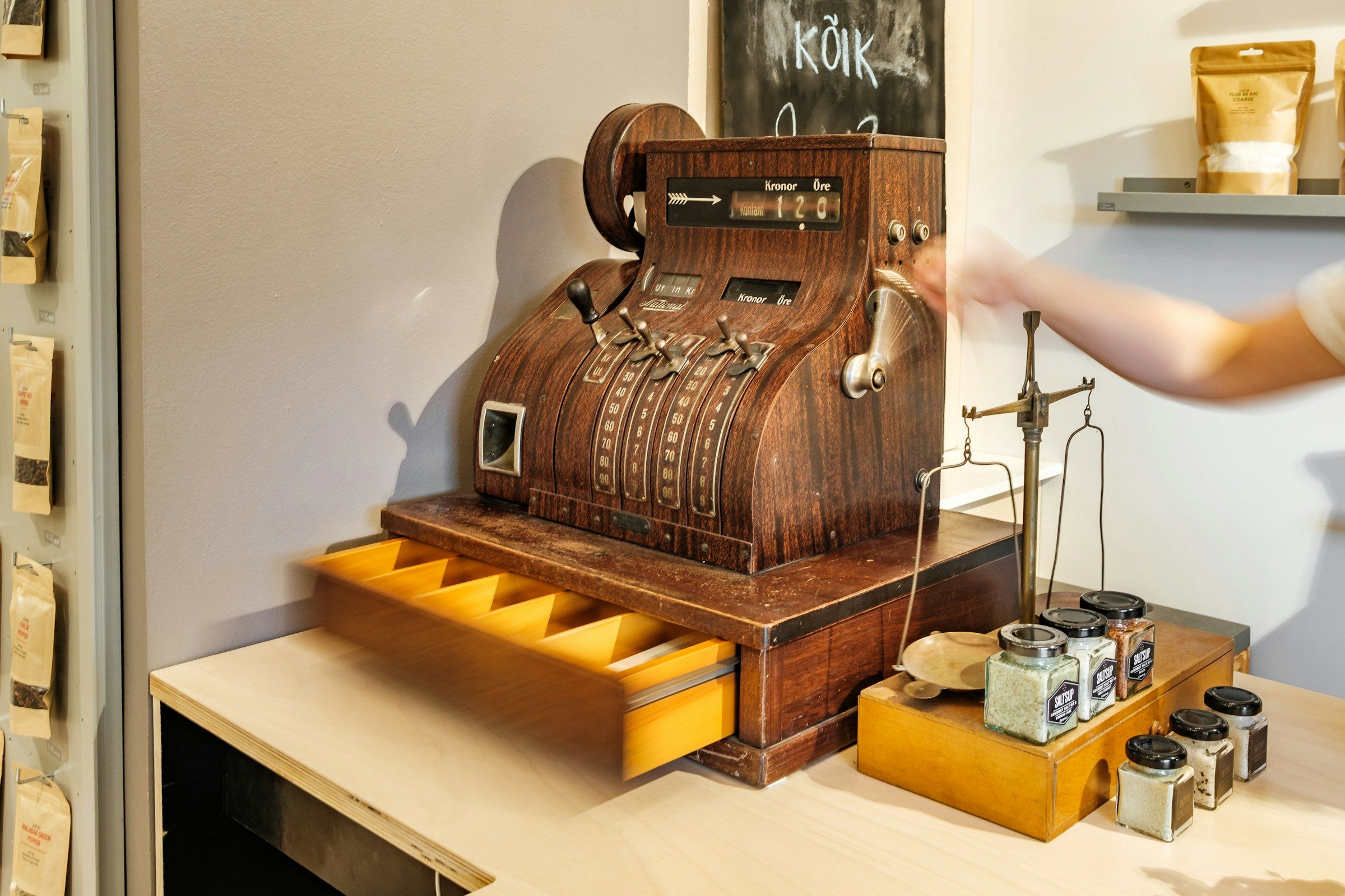 A wooden antique cash register with its drawer open on a small shop counter, beside brass scales and jars; an arm reaches in from the right edge