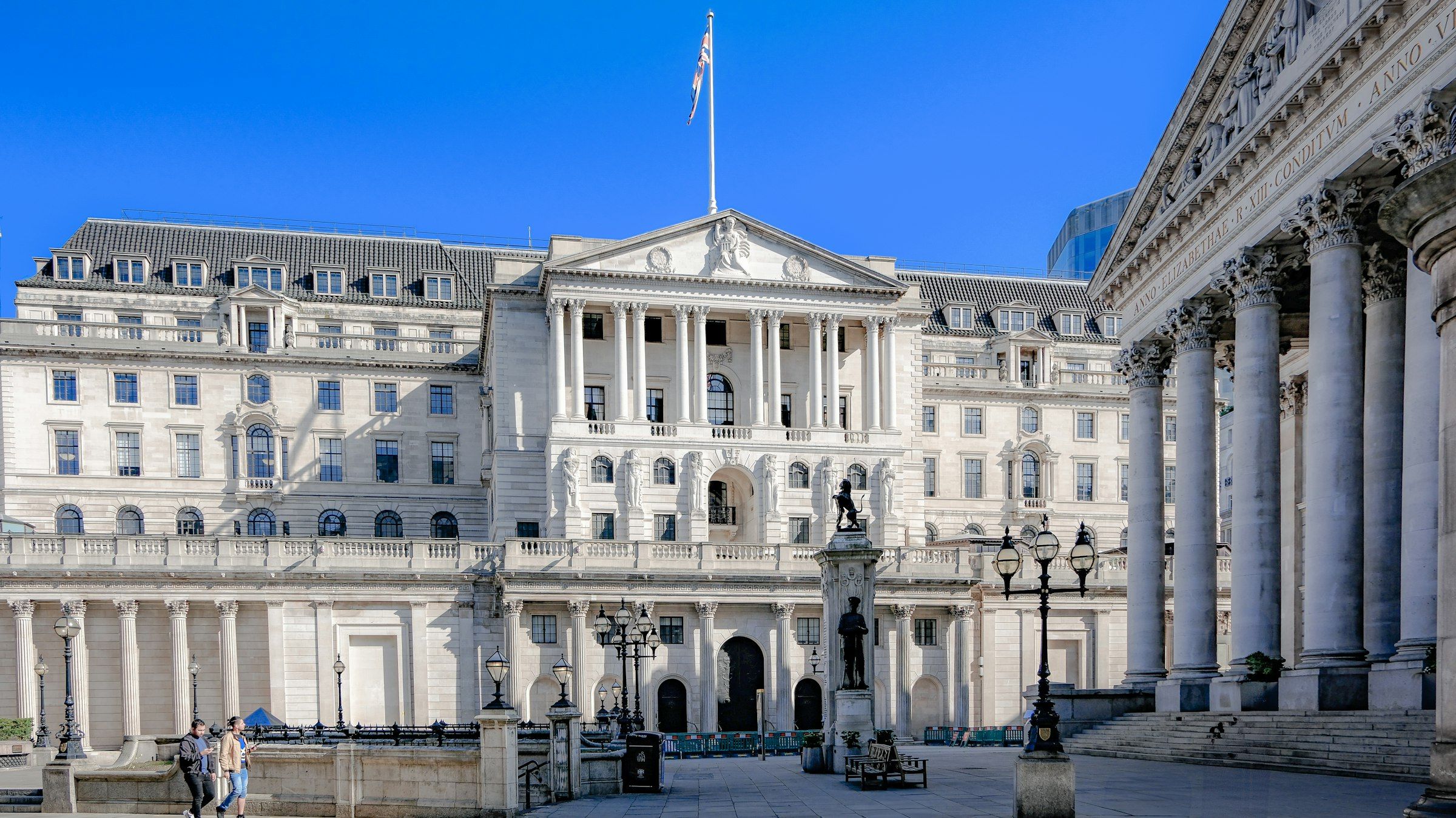 The neoclassical stone facade of the Bank of England under a clear blue sky, with columned porticos, street lamps and a flag on the roofline