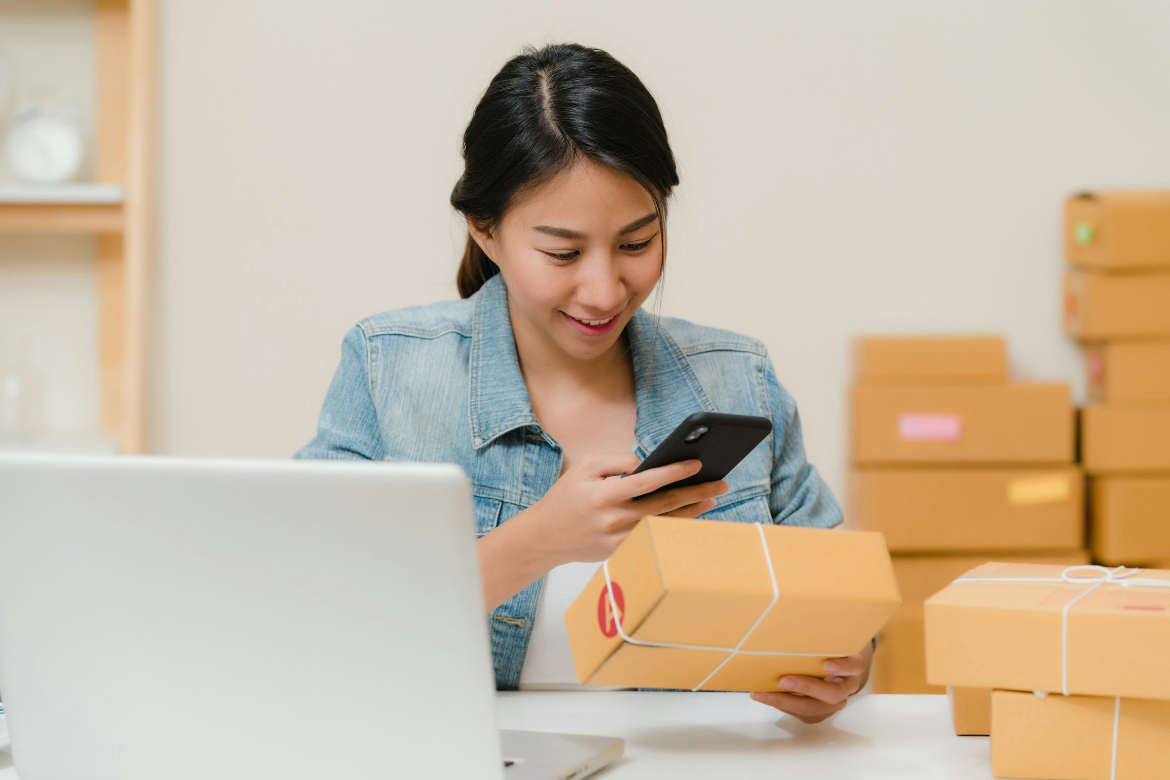 A merchant checking a package against her phone beside a laptop and stacked boxes