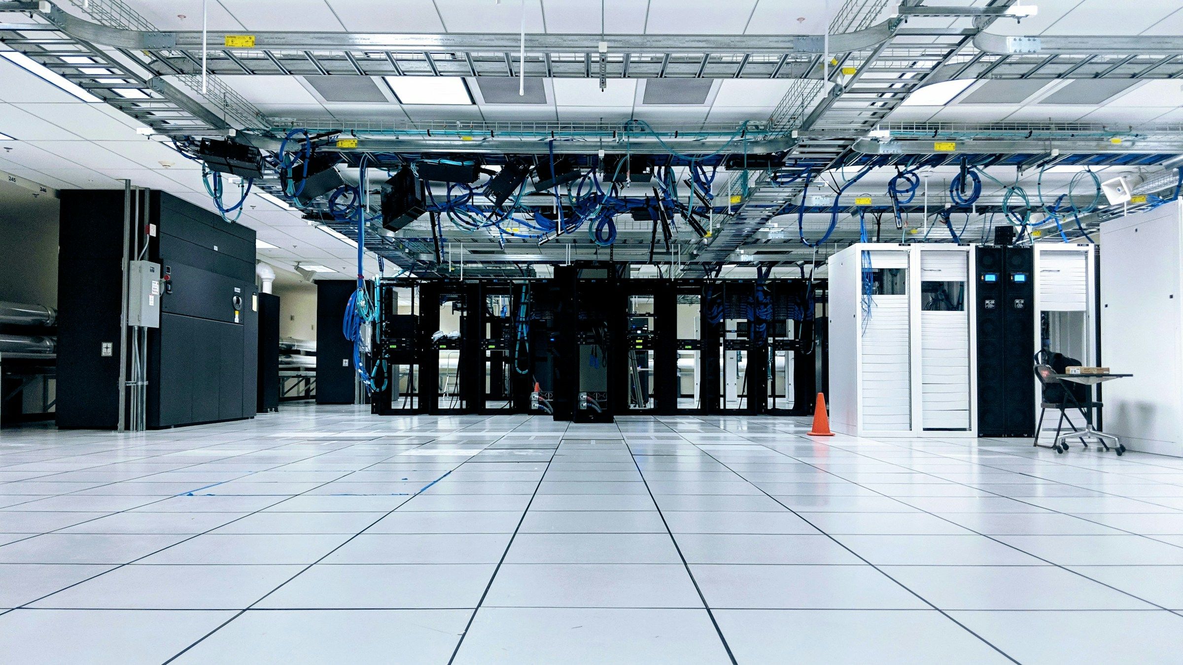 A data centre hall with rows of black and white cabinets under blue overhead cabling
