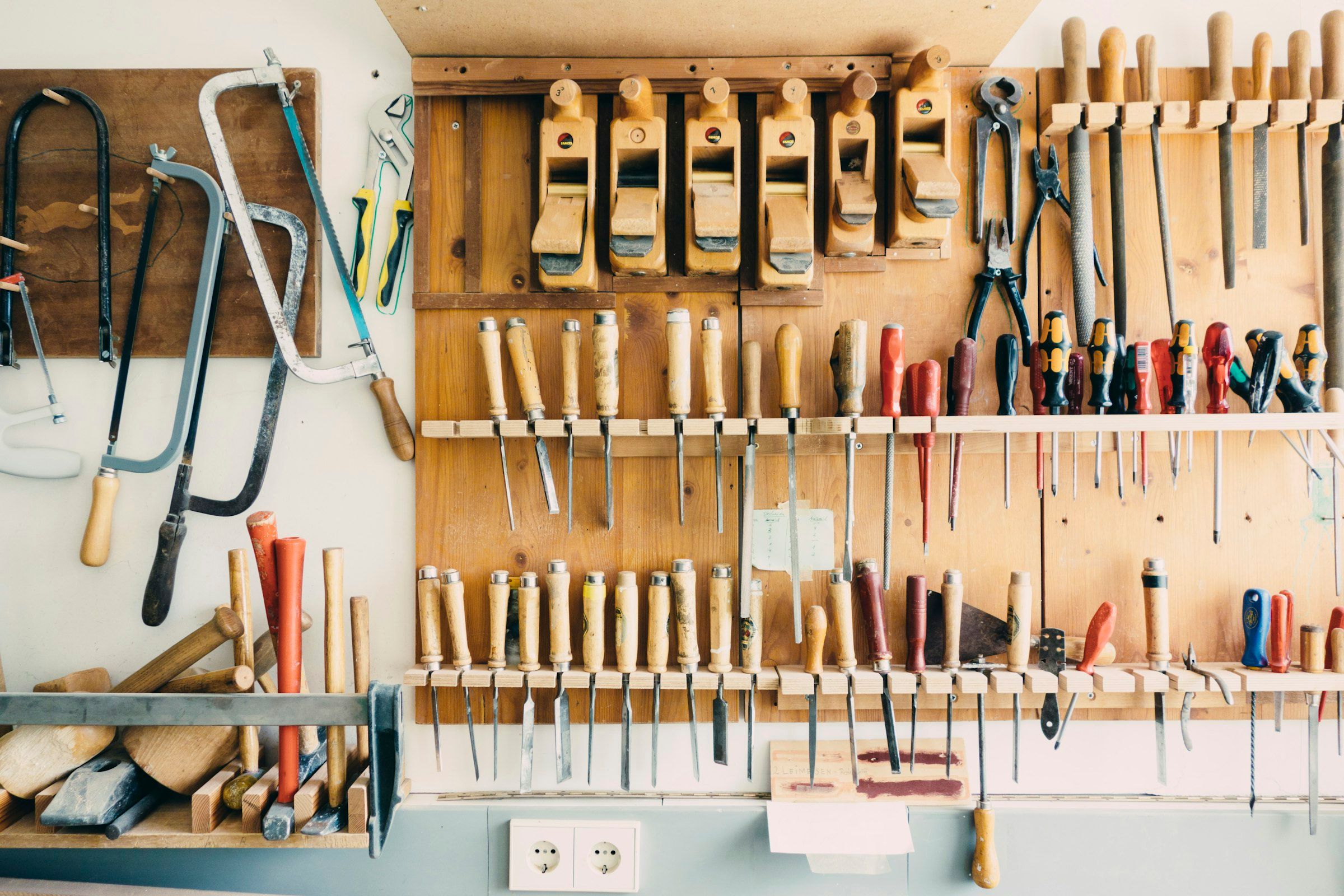 A workshop wall rack holding rows of chisels, hand saws, screwdrivers and wooden planes above a workbench