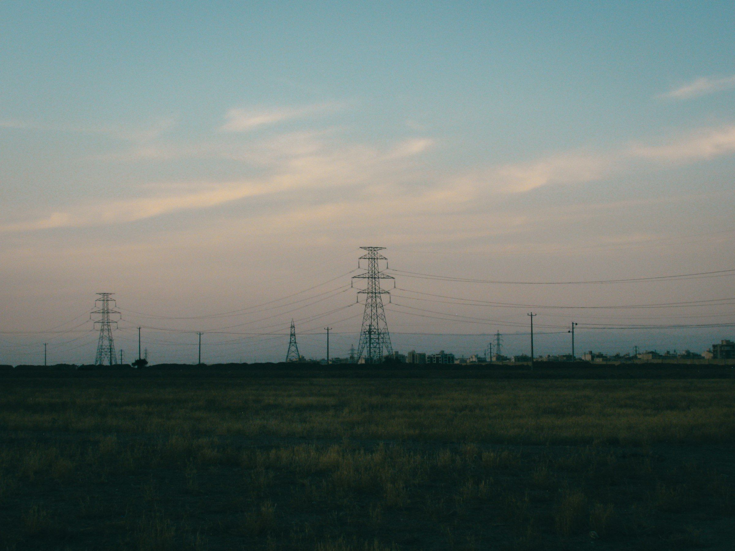 Lattice transmission towers and power lines crossing a flat, empty field at dusk, with a low settlement on the horizon