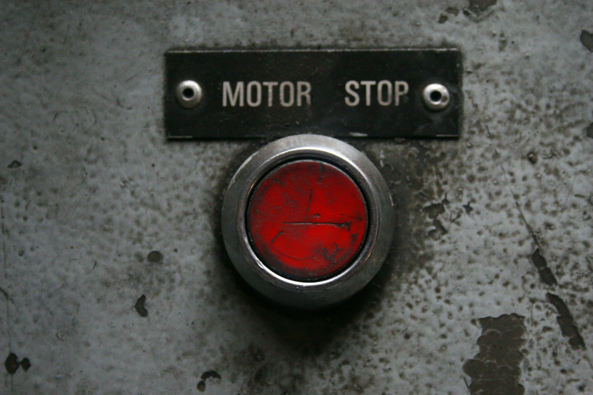 A worn red push button set into scratched grey metal, beneath a small engraved plate reading MOTOR STOP