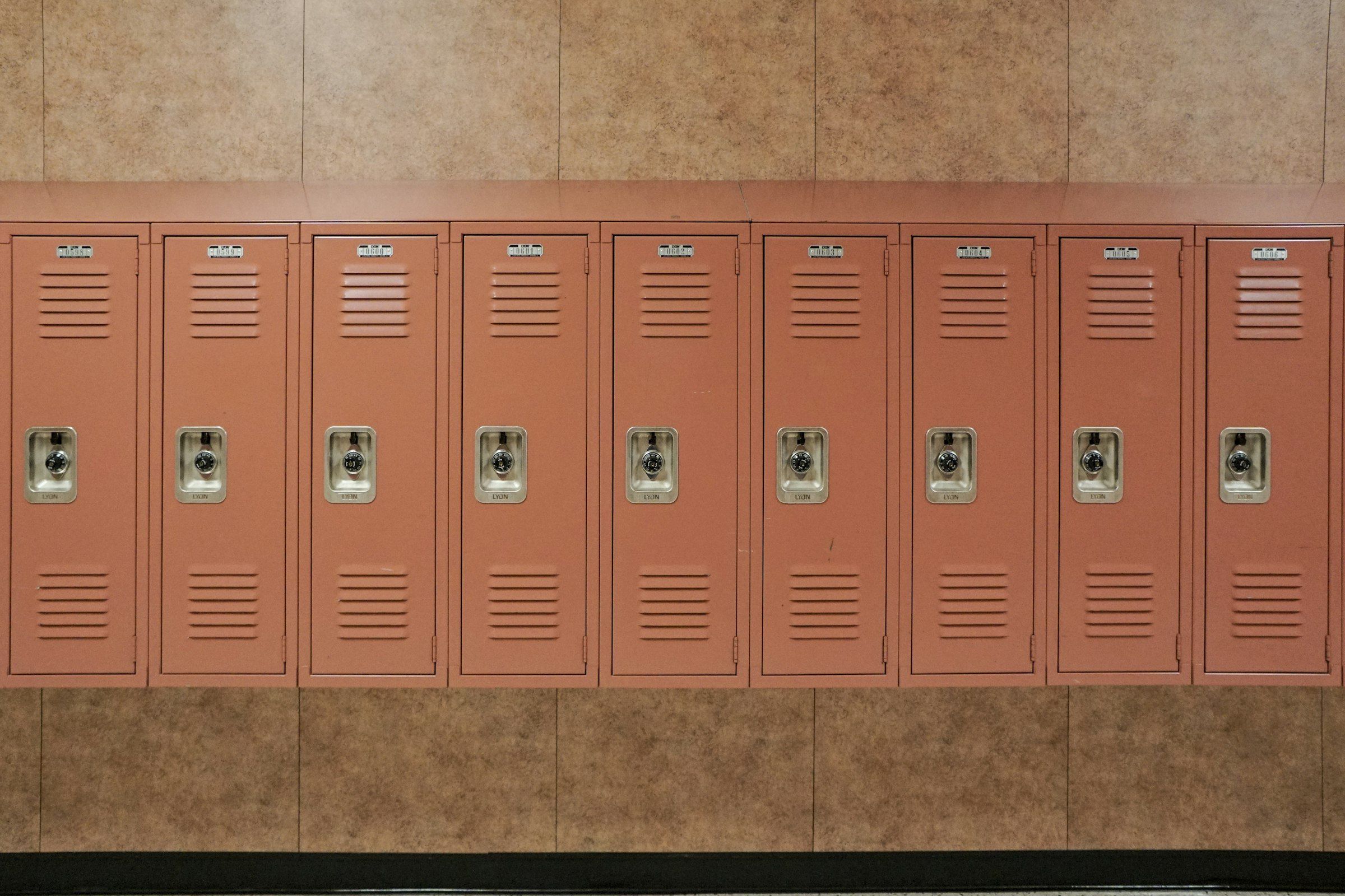 A row of closed salmon-coloured metal school lockers with combination dials, against a tiled corridor wall