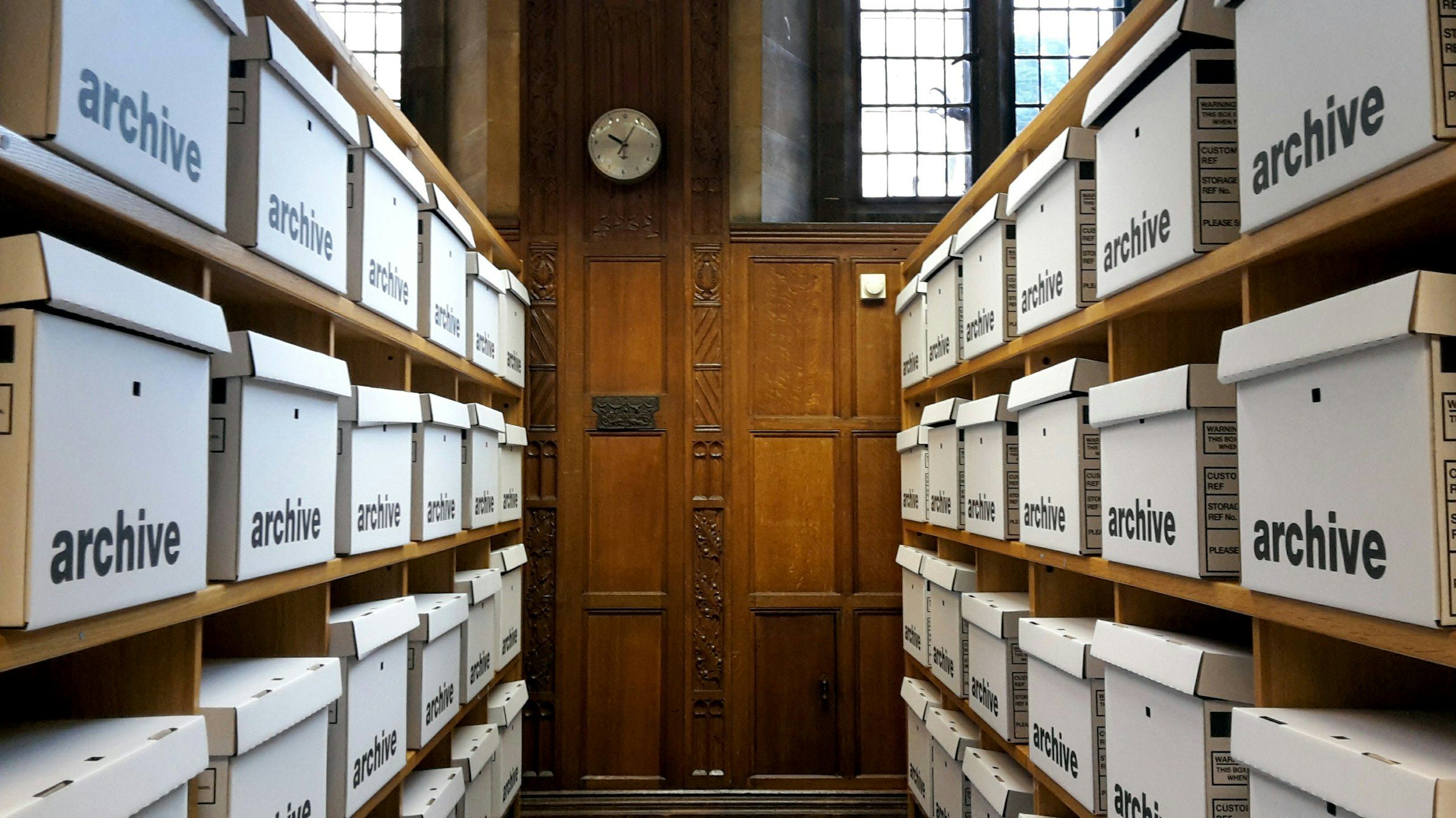 Two facing shelf stacks of white storage boxes each printed with the word archive, forming an aisle that ends at a closed wooden door beneath a clock