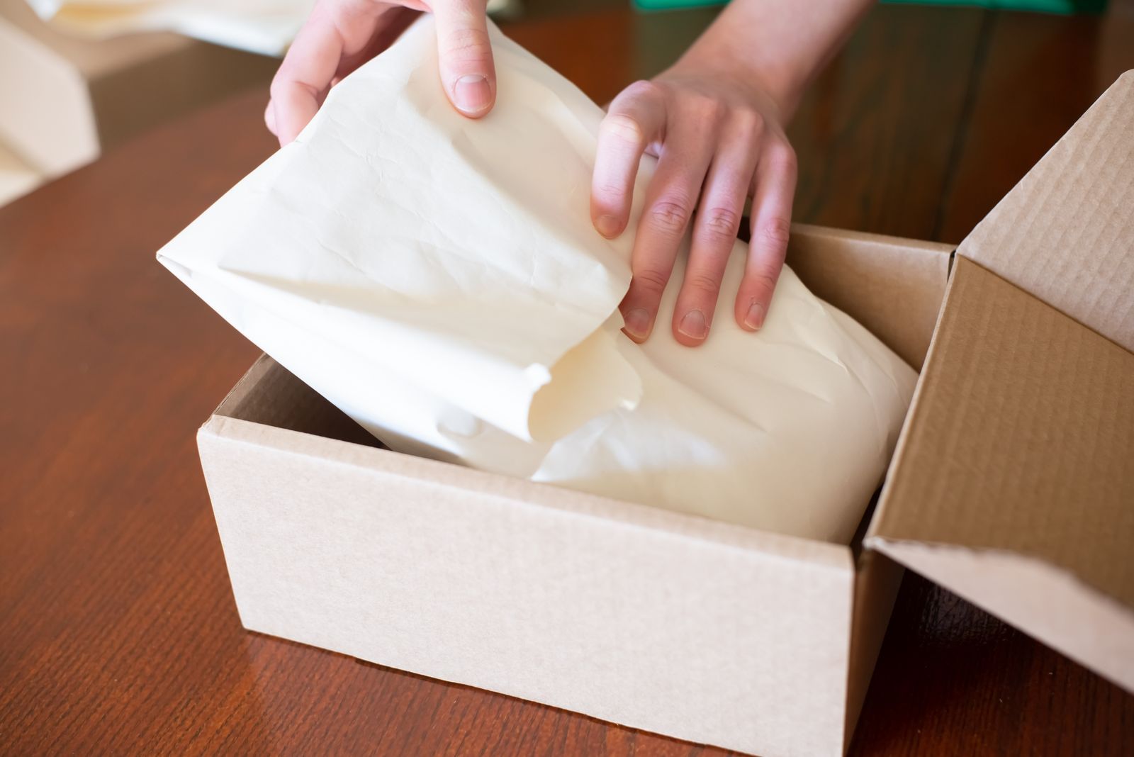 Hands placing a wrapped item into an open cardboard box on a wooden table