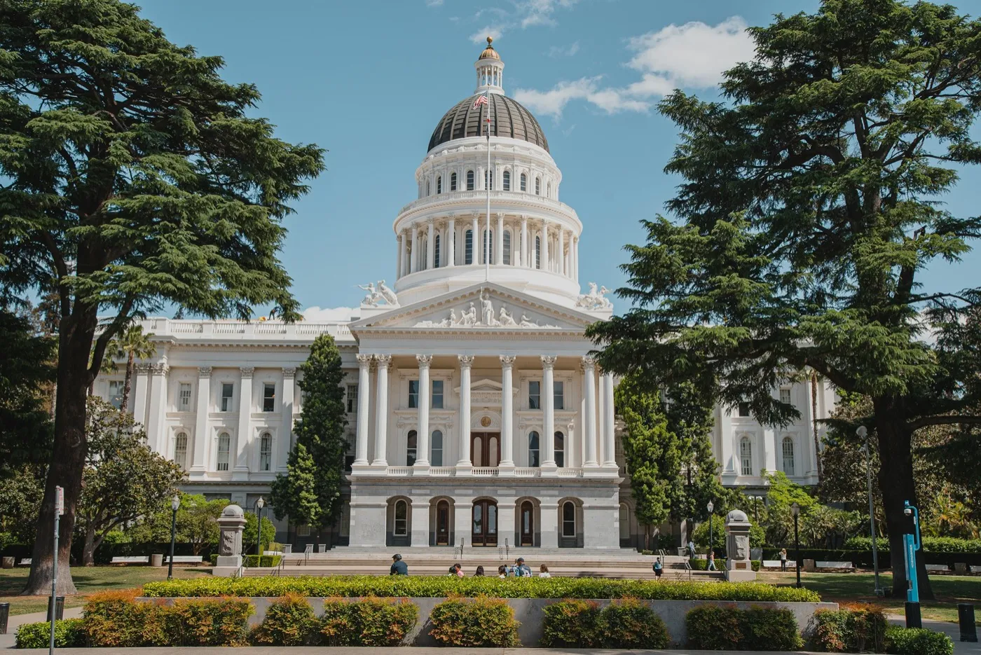 The California State Capitol in Sacramento, its dome and columned portico framed by trees, with flower beds and a few people on the lawn in front