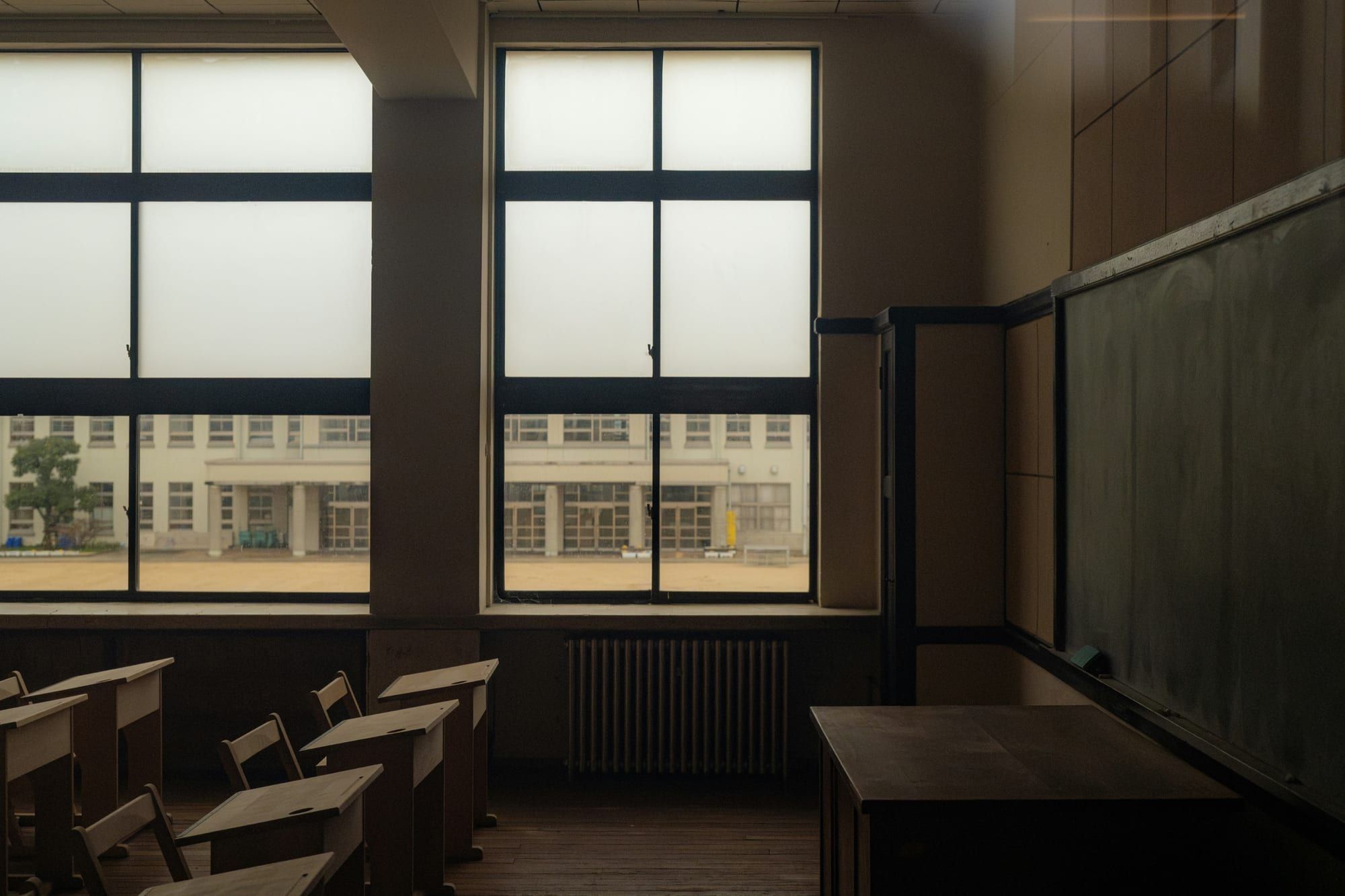 Rows of empty wooden school desks beside tall windows, with a blank chalkboard on the right wall