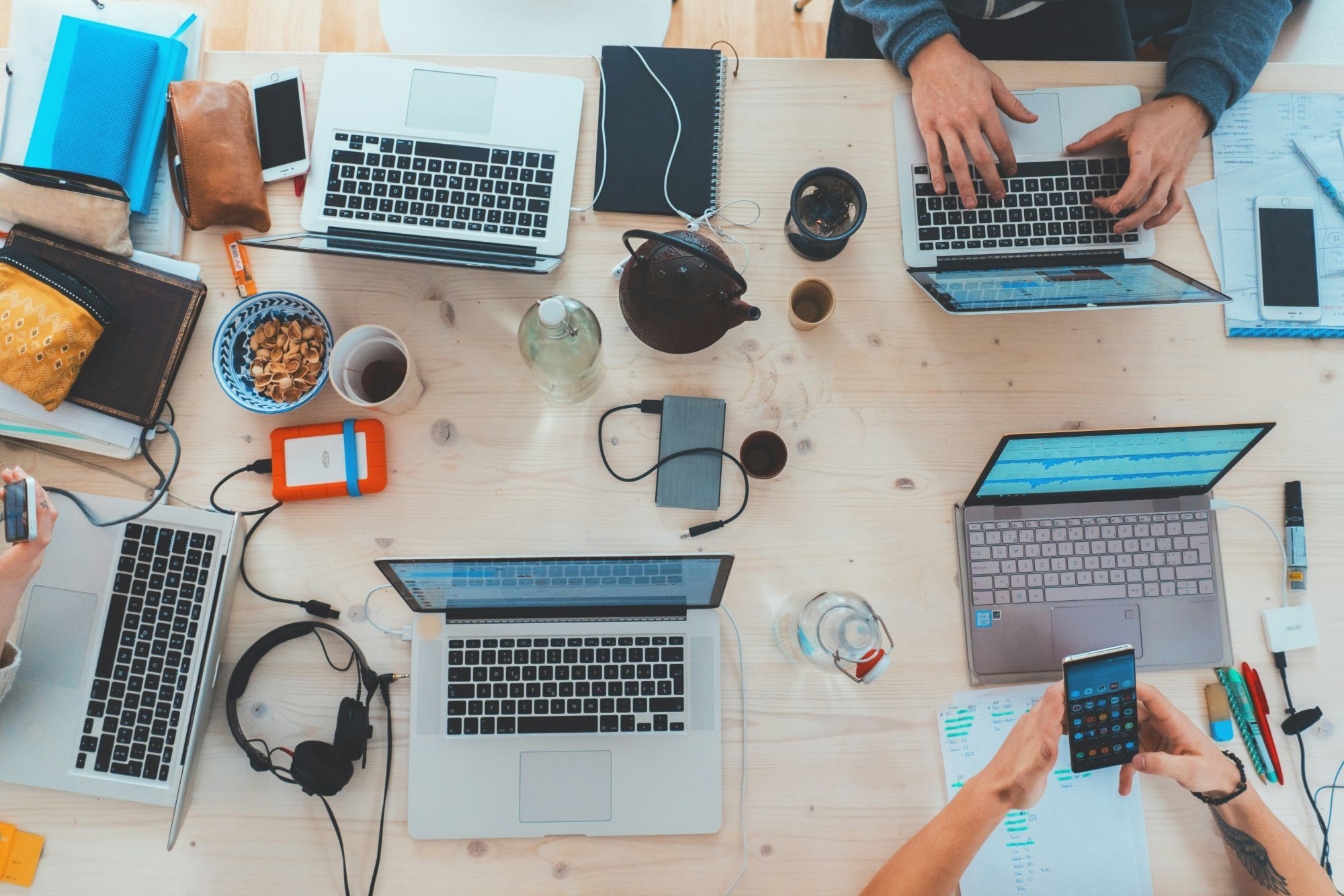 Overhead view of a shared wooden table with five open laptops, phones, headphones, a teapot and a bowl of pistachio shells, with people typing and holding a phone at the edges