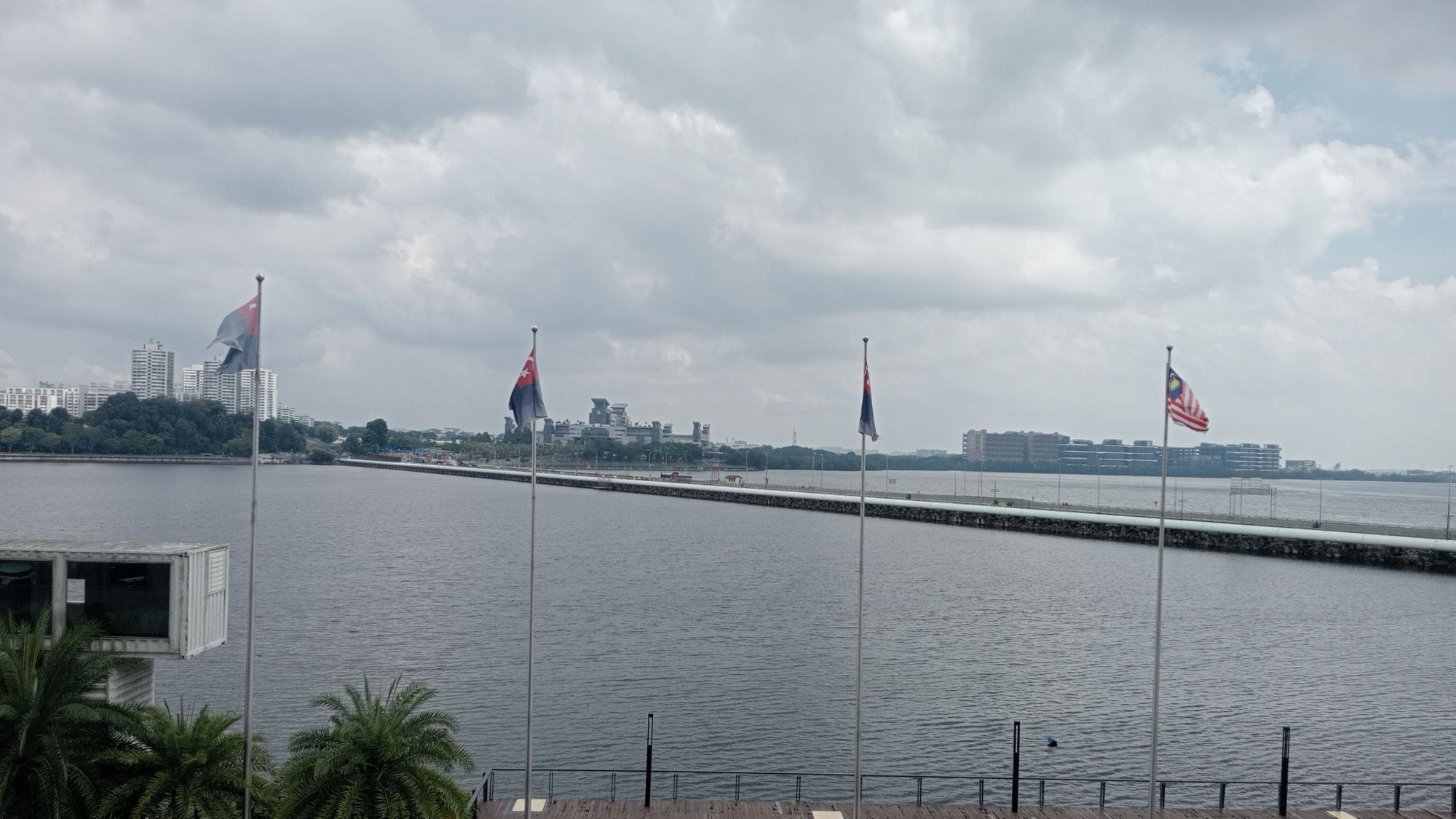 Flags, including Malaysia's, on poles above a waterfront deck, with the causeway running across the Johor Strait to buildings on the far shore under a cloudy sky