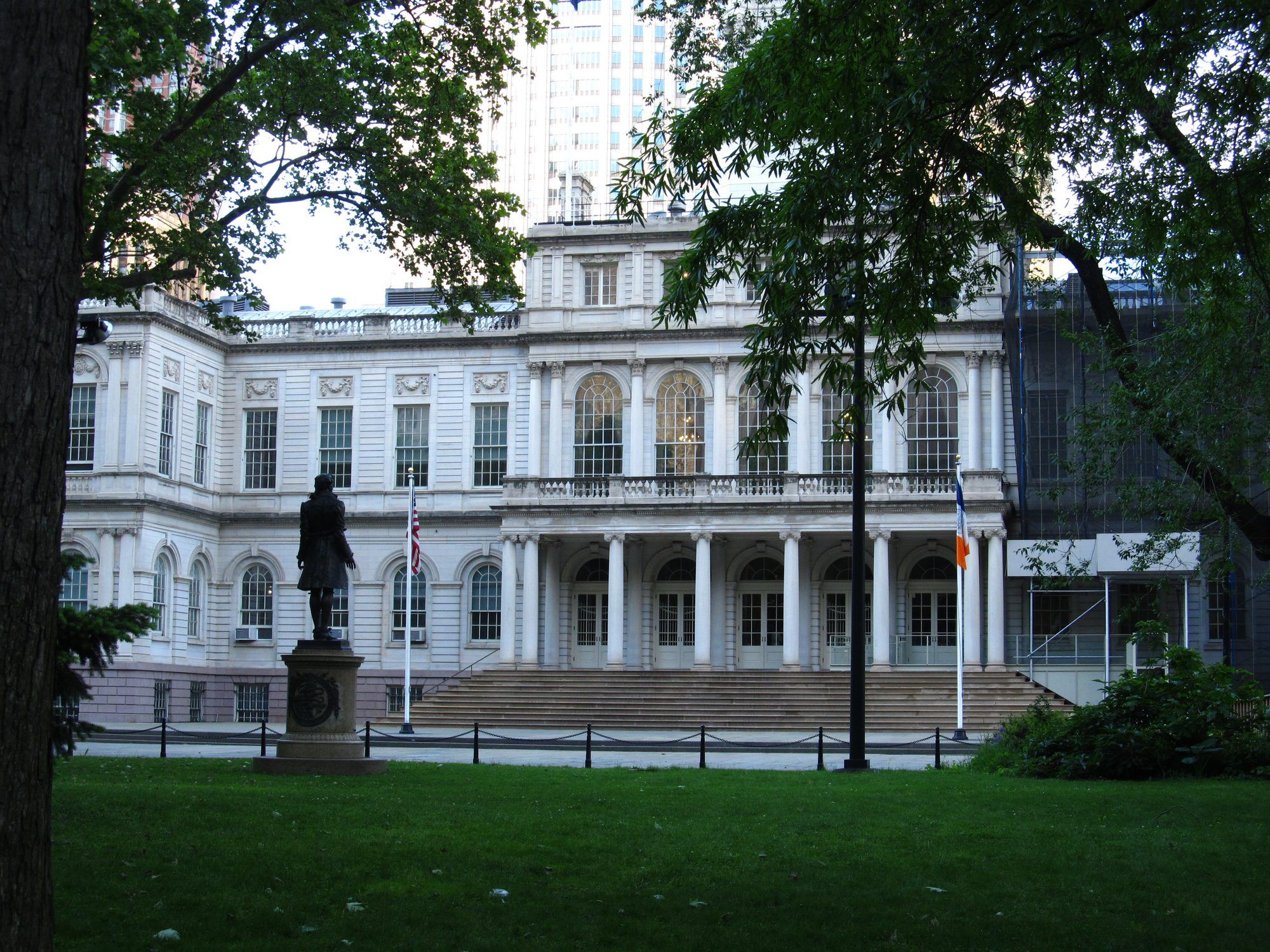 The white columned front of New York City Hall seen across a lawn through trees, with a bronze statue on a stone plinth, the US flag and the New York City flag beside the entrance steps