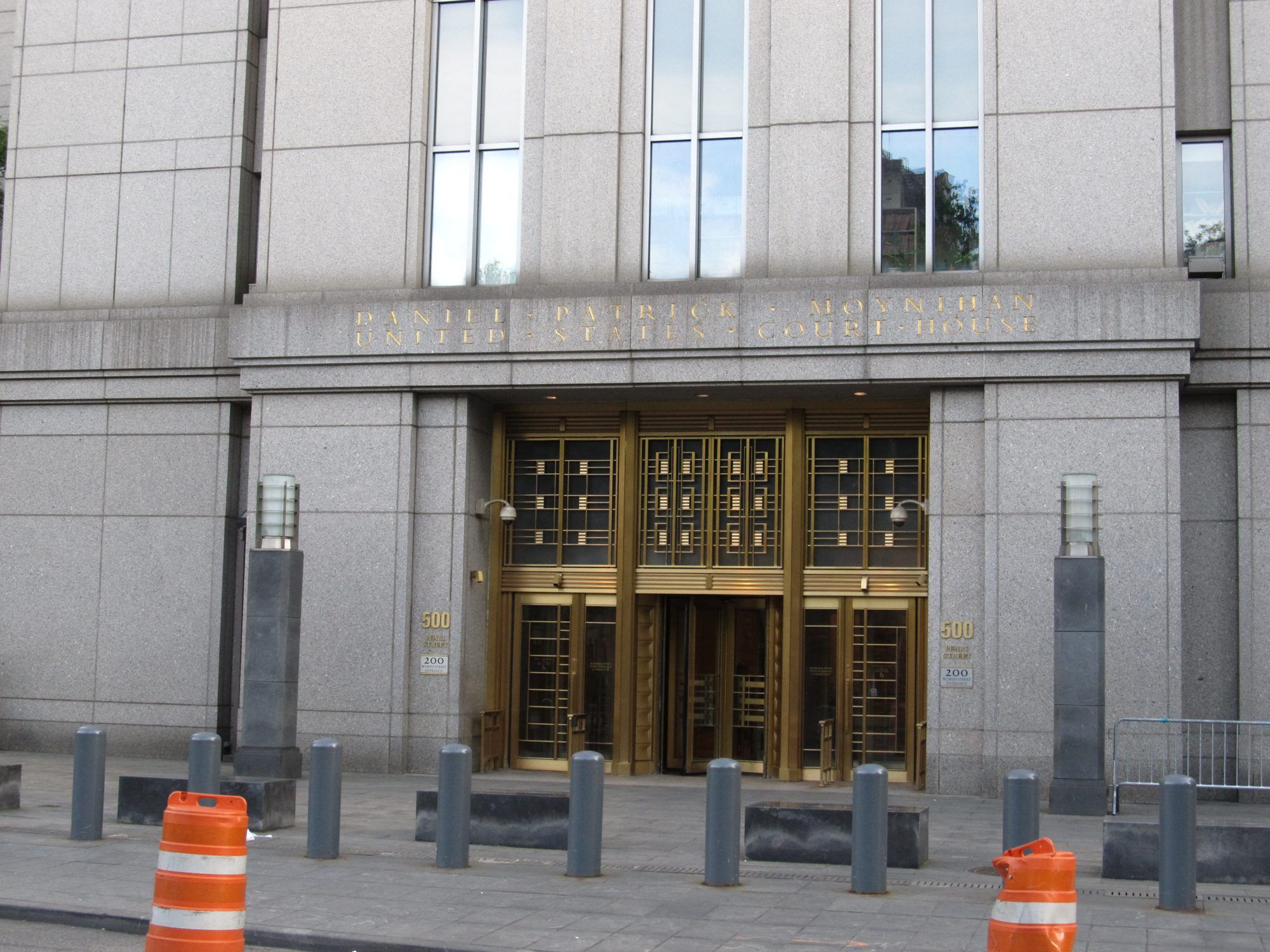 The bronze-doored entrance of the Daniel Patrick Moynihan United States Courthouse in Manhattan, with bollards and orange traffic barrels on the pavement