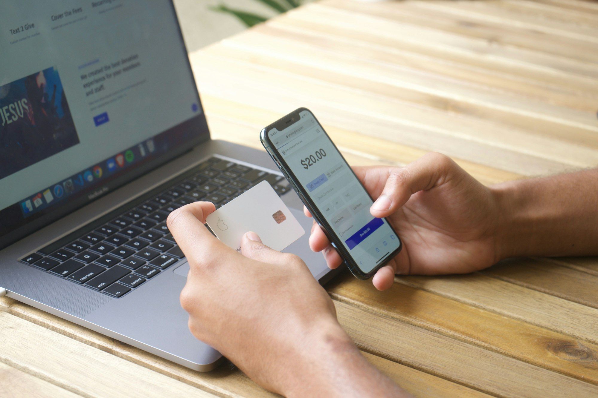 A person holding a payment card in one hand and a smartphone in the other above an open laptop on a wooden table