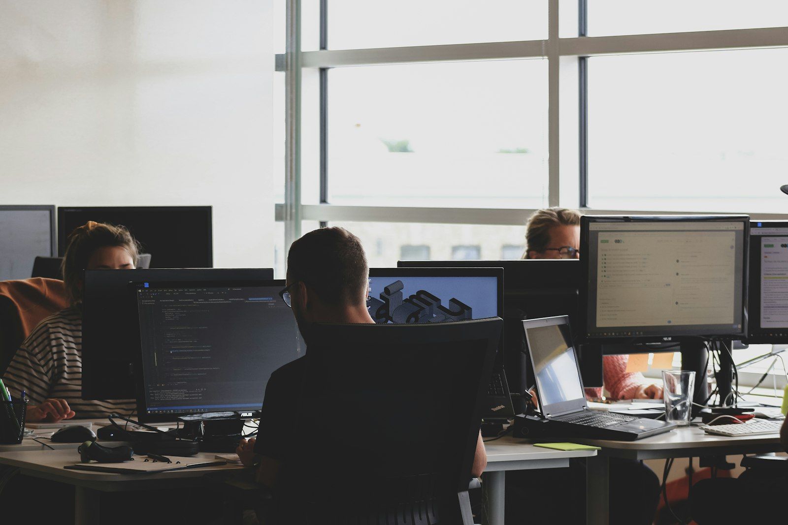 Three people working at desks with multiple monitors in a bright open-plan office