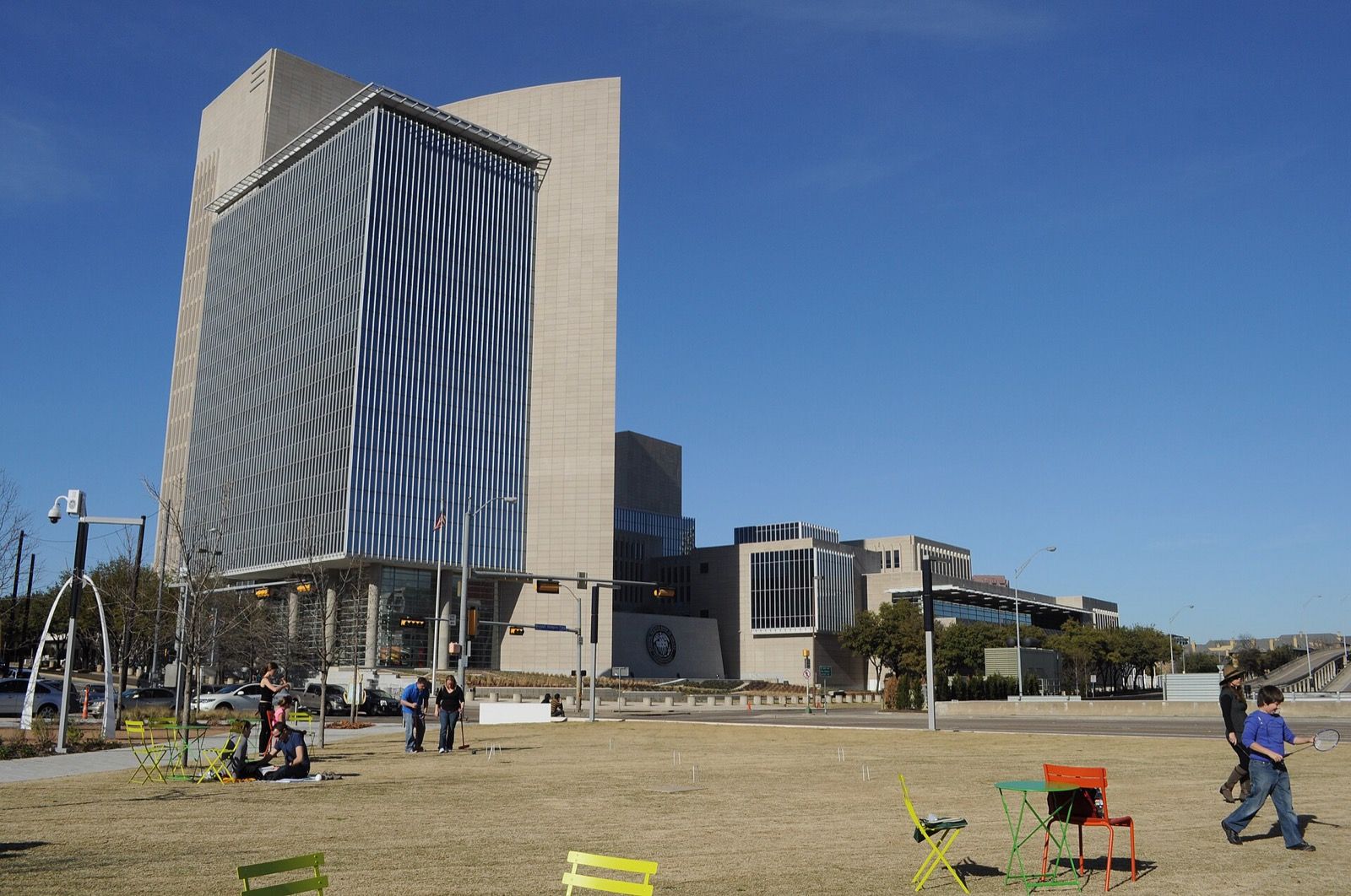 The Federal Reserve Bank of Dallas building seen from Klyde Warren Park under a clear sky, with people on the lawn and folding chairs in the foreground