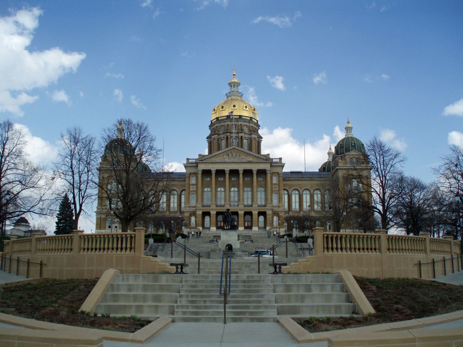 The Iowa State Capitol in Des Moines, its gold central dome above a columned portico, seen from the foot of a wide stone staircase under a partly cloudy sky