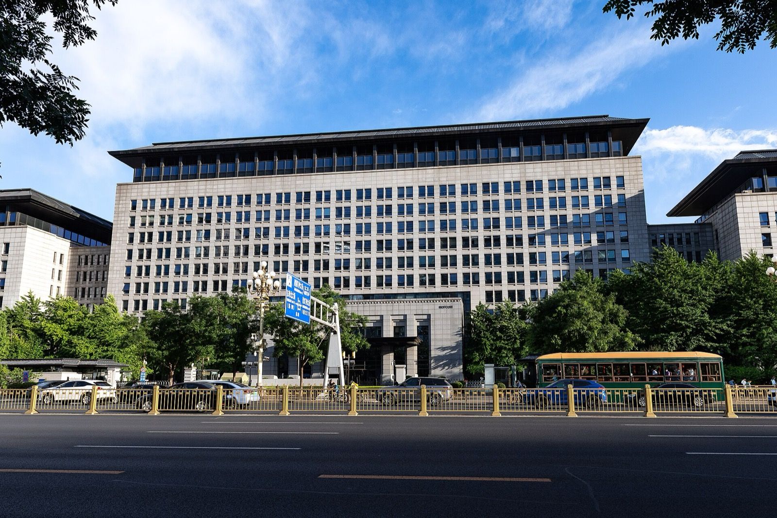 The Ministry of Commerce building in central Beijing, a long stone-clad block behind trees and a yellow roadside railing, with a green bus passing under a blue sky with scattered cloud