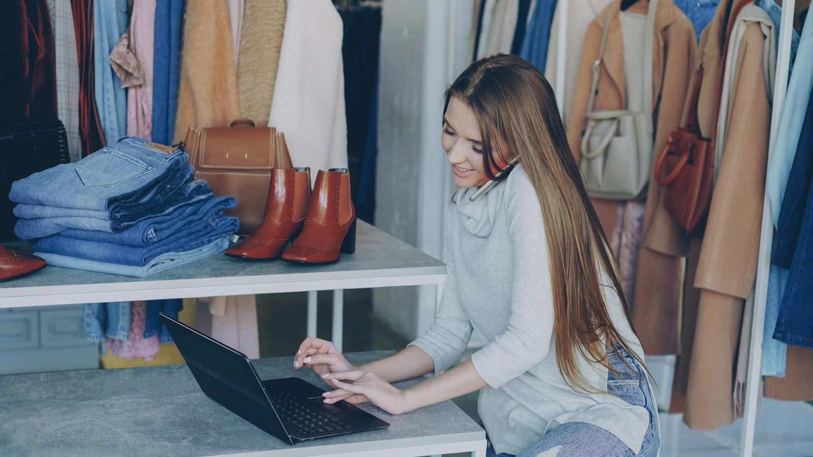 A woman on a phone call works at a laptop in a clothing shop, with folded jeans and boots on the shelves and bags around her