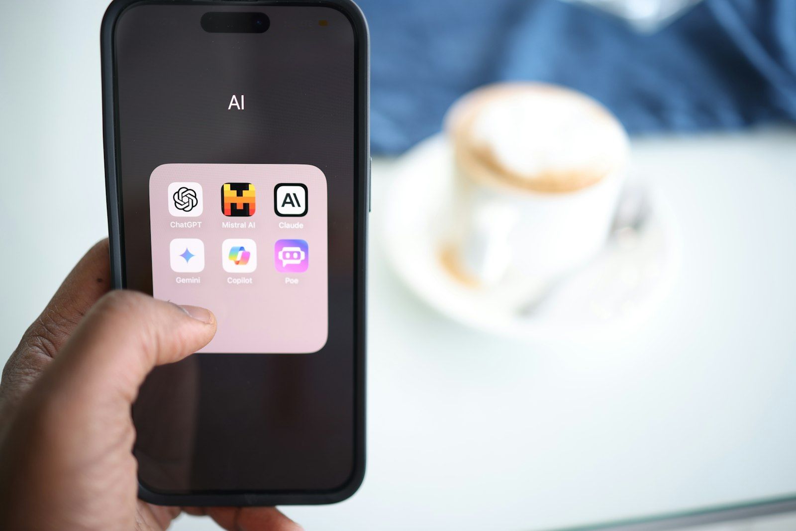 A hand holds a phone showing a folder labelled AI with app icons for ChatGPT, Mistral AI, Claude, Gemini, Copilot and Poe, with a cup of coffee blurred in the background
