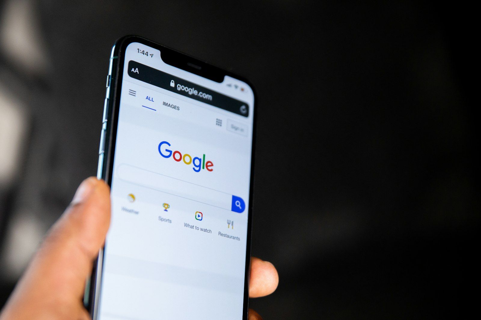A hand holding a smartphone showing the Google search page at google.com, against a dark background
