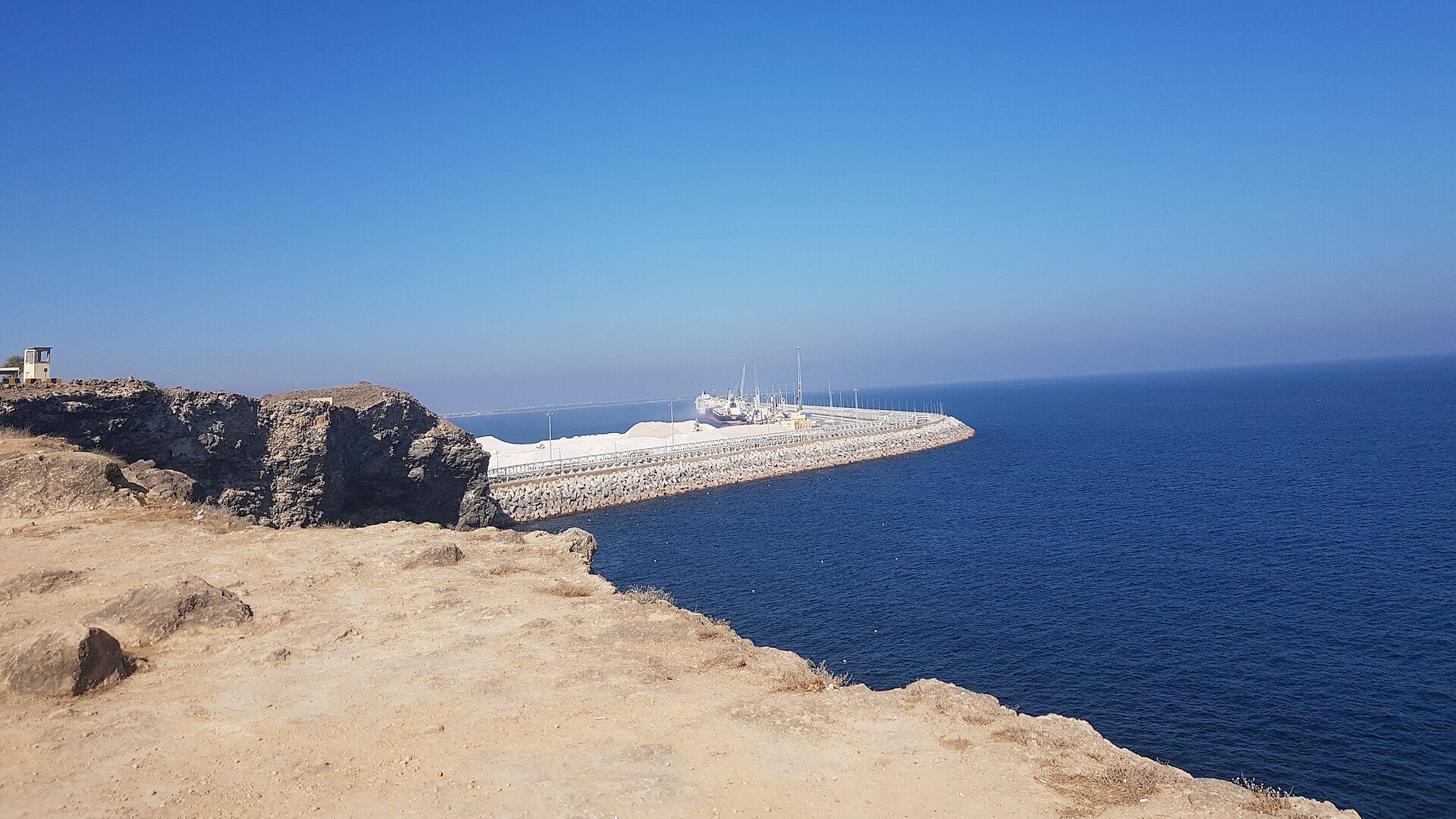 The breakwater of Salalah's port in Oman, with ships and cranes at the quay, seen from a rocky clifftop above the Arabian Sea